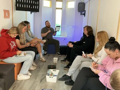 Ein Erzieher sitzt vor Kopf im Innenraum der Tiny Church, umringt von Jugendlichen und Erwachsenen. Er zeigt ihnen ein Bild, während sie selbst Bilder auf ihrem Schoss ausmalen.