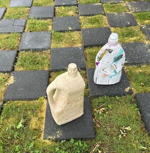 Zwei alte Waschmittelflaschen, beklebt mit Pappmache, stehen auf einem Schachbrett auf einer Wiese.
