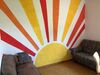 Ein Zimmer zeigt ein lebhaftes Sonnenbild mit roten, orangefarbenen und gelben Strahlen an der Wand, davor stehen eine braune Couch und eine Couch mit floralem Muster.